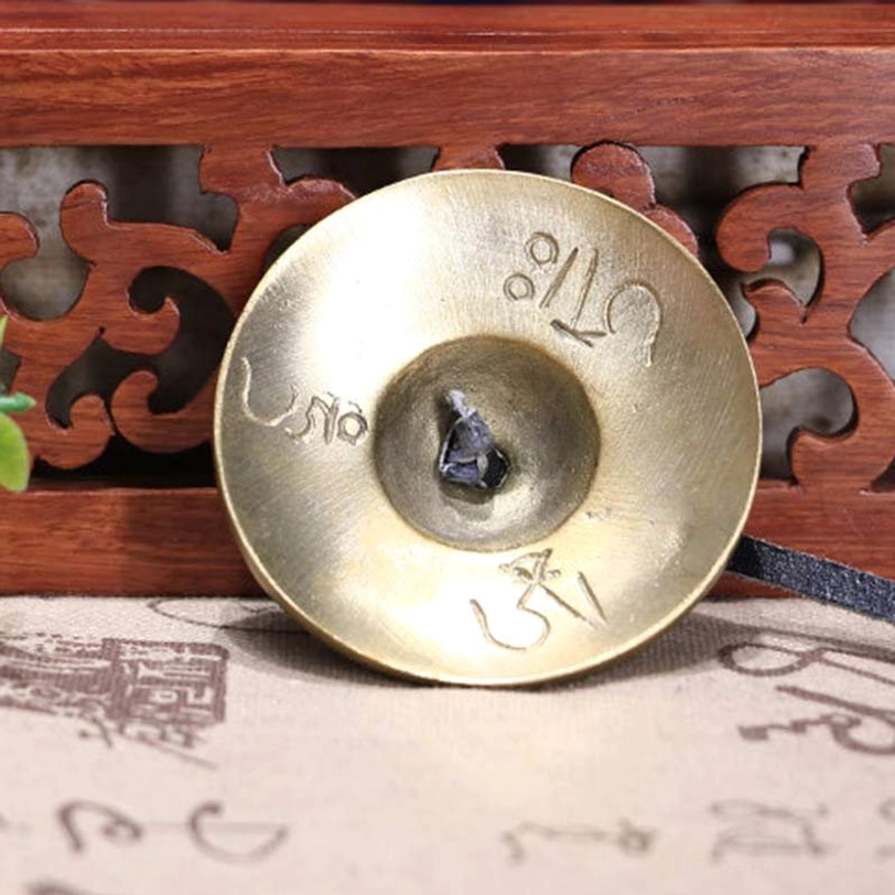 Single tibetan tingsha bell close-up showing engraved brass surface with sacred symbols and leather attachment on fabric backdrop