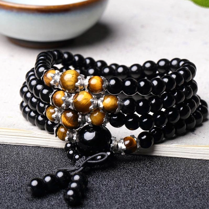 Stacked obsidian and tiger eye mala bracelets on dark table with open book, showing glossy bead detail