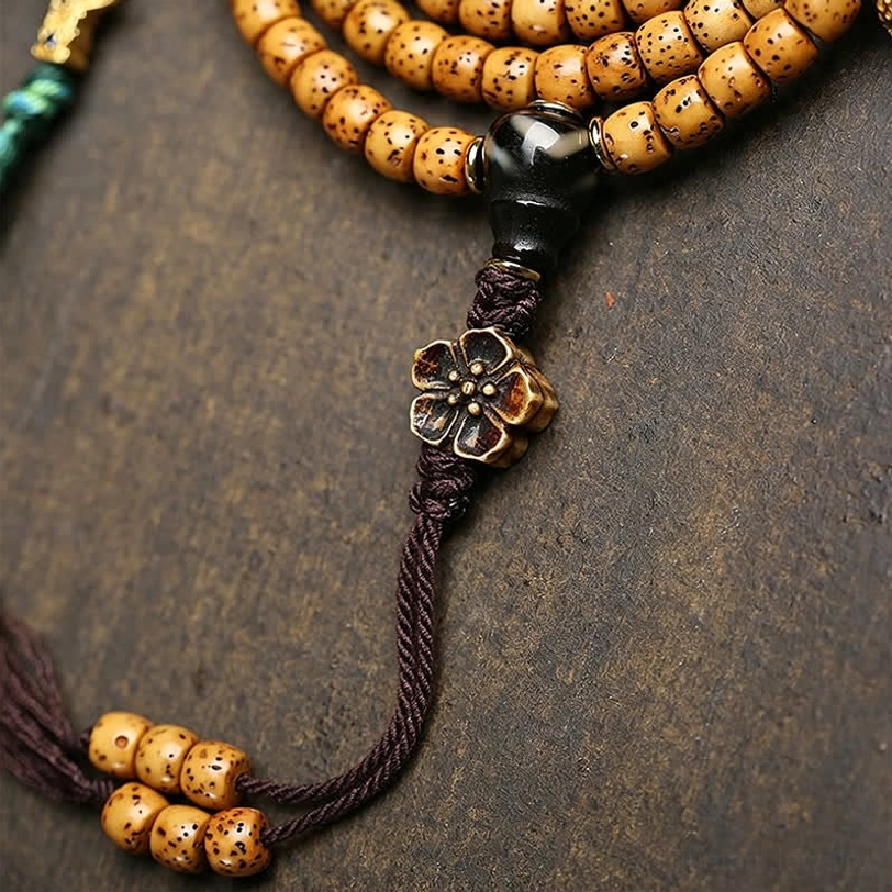 Angled close-up of Star Moon Bodhi mala beads showing tan cylinder beads black spacer bead ornate metal flower knot and braided cord detail