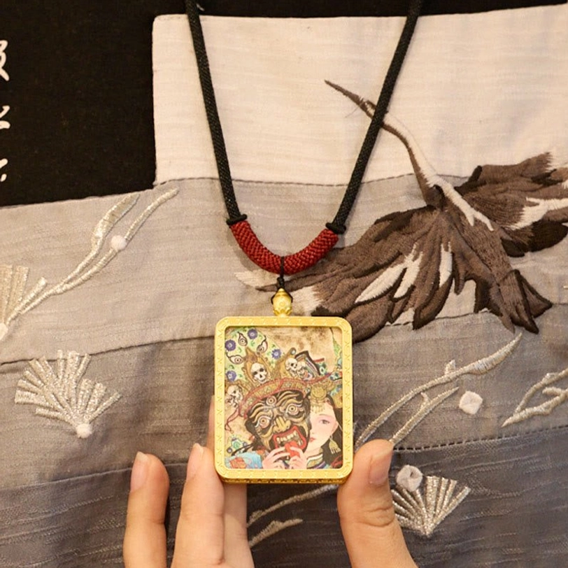 Thangka pendant necklace displayed on embroidered traditional Tibetan fabric, showing dual figure artwork and red-black woven cord