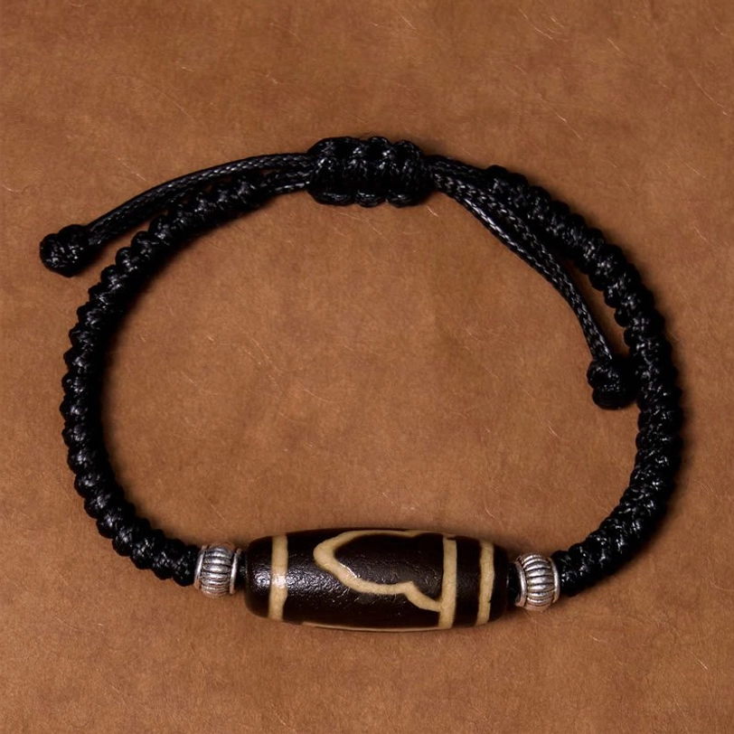 Three-eye dzi bead bracelet detail shot showing sacred three-eye pattern with braided black rope on neutral background