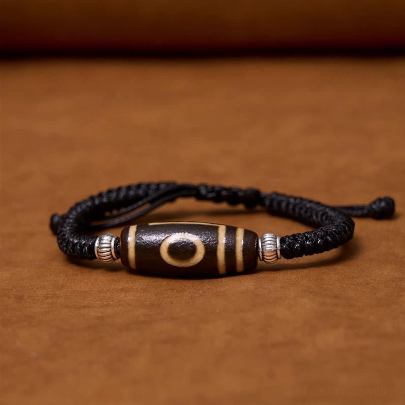 Tibetan three-eye dzi bead bracelet flat side view on brown wooden table with black braided cord and silver spacers