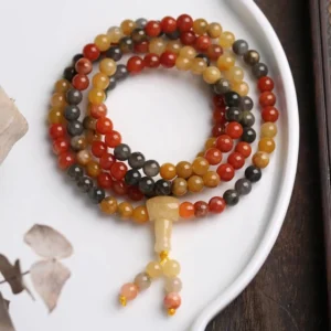 108-bead natural red agate mala bracelet with yellow agate and black onyx beads displayed on white ceramic dish over wooden table, showcasing polished gemstones with carved guru bead and silk tassel pendant