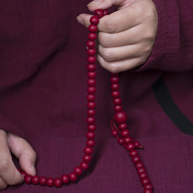 108 Abelia Biflora mala beads prayer bracelet with red wooden beads and tassel pendant held in hands against traditional maroon tibetan robe background