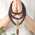 108 mala beads bracelet with three-eye dzi bead held in hand showing deep brown wooden beads, light spacers, and patterned dzi charm