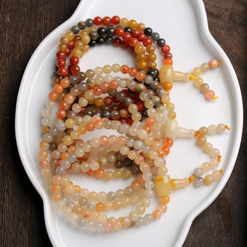 Multiple sets of handmade 108-bead agate mala bracelets displayed on white ceramic dish, showing various warm color tones of natural red agate and yellow agate stones with matching silk tassels