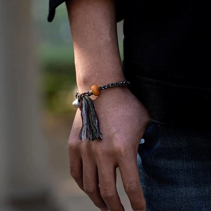 Tibetan amber bracelet styled on wrist outdoors showing dark braided strap, natural amber stone, silver charm, and protective tassel