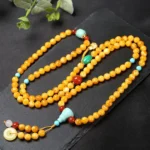 Top-down view of 108-bead Tibetan amber mala with turquoise and red agate accents, peace buckle charm and metal guru bead on dark slate