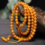 Tibetan amber 108 bead mala close-up with warm orange polished beads, dark red spacer, and yellow tassel on wooden display