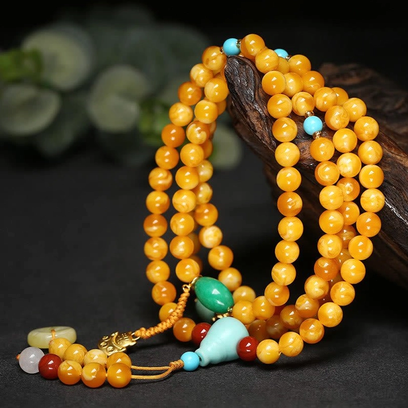 Close-up of smooth round amber mala beads with tassel pendant and gemstone decorations on wooden surface with green foliage