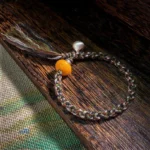 Tibetan amber and sterling silver steelyard weight protection bracelet flat lay on wooden mat showing multicolor braided cord, golden amber bead, and silver charm with tassel