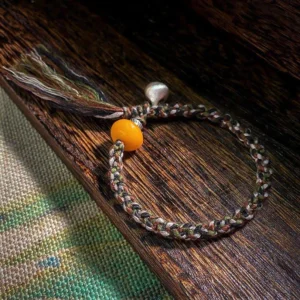 Tibetan amber and sterling silver steelyard weight protection bracelet flat lay on wooden mat showing multicolor braided cord, golden amber bead, and silver charm with tassel
