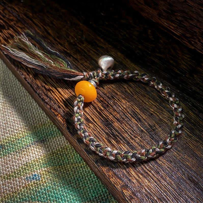 Tibetan amber and sterling silver steelyard weight protection bracelet flat lay on wooden mat showing multicolor braided cord, golden amber bead, and silver charm with tassel