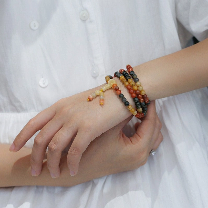 Multiple handmade 108-bead agate mala bracelets worn on single wrist with white shirt background, close-up hand view showcasing stacked styling with various warm-tone gemstones and tassel pendants