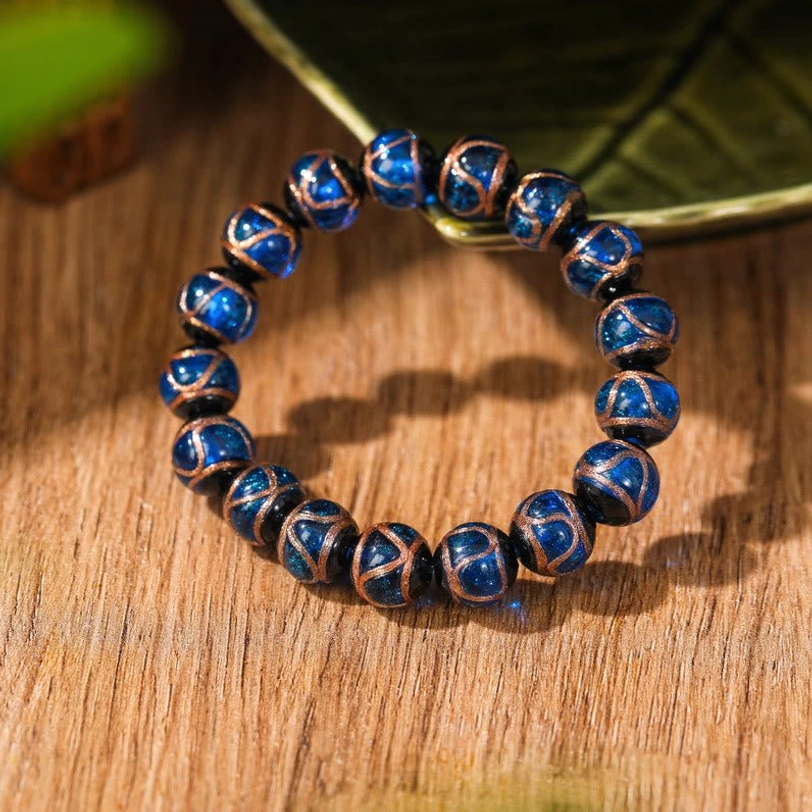 Single blue-toned Tibetan liuli glass bead bracelet with deep blue glazed beads and gold decorative stripes on wooden table with green leaf accent