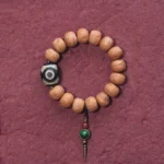 Tibetan bodhi seed dzi bead mala bracelet overhead view with three-eyed dzi focal bead and brass charm on wine-red background