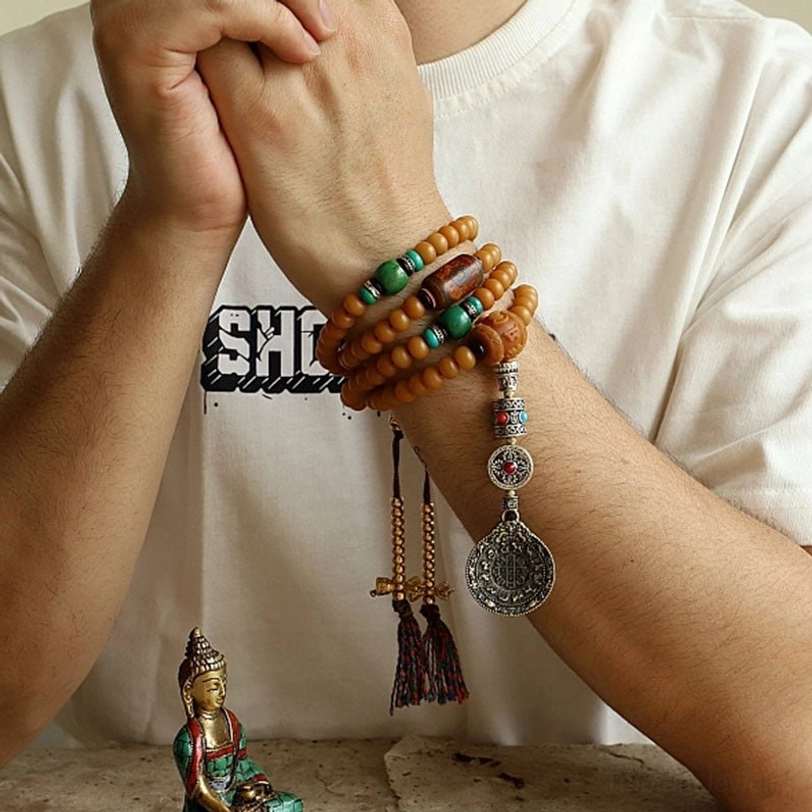 Multi-wrap Tibetan bodhi seed mala bracelet on wrist showing stacked tan beads, silver and stone embellishments, hanging tassels, and Bagua charm