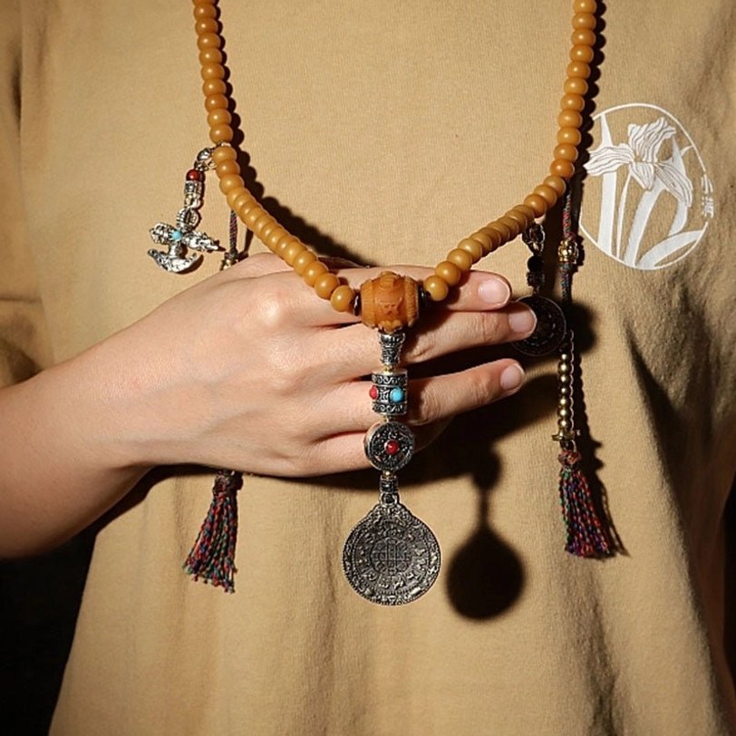Tibetan bodhi seed mala necklace worn on hand showing tan bead strand, silver decorative dangles, hanging tassels, and large Bagua pendant close-up