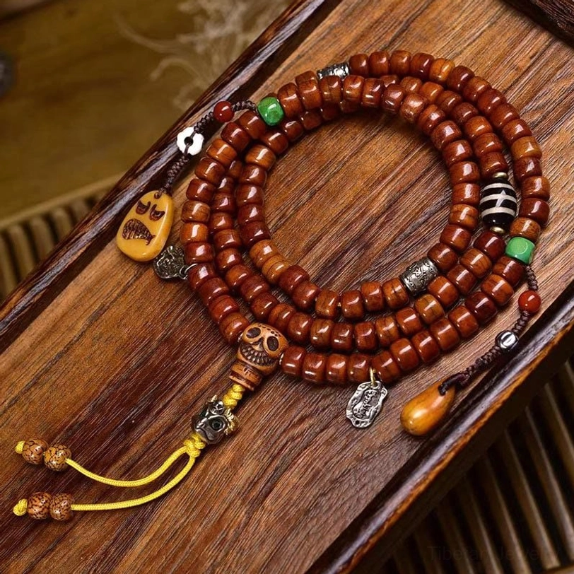 Tibetan 108 bone bead mala with hand-painted dzi beads flat lay on wooden surface showing reddish-brown barrel beads and skull guru bead with yellow tassel