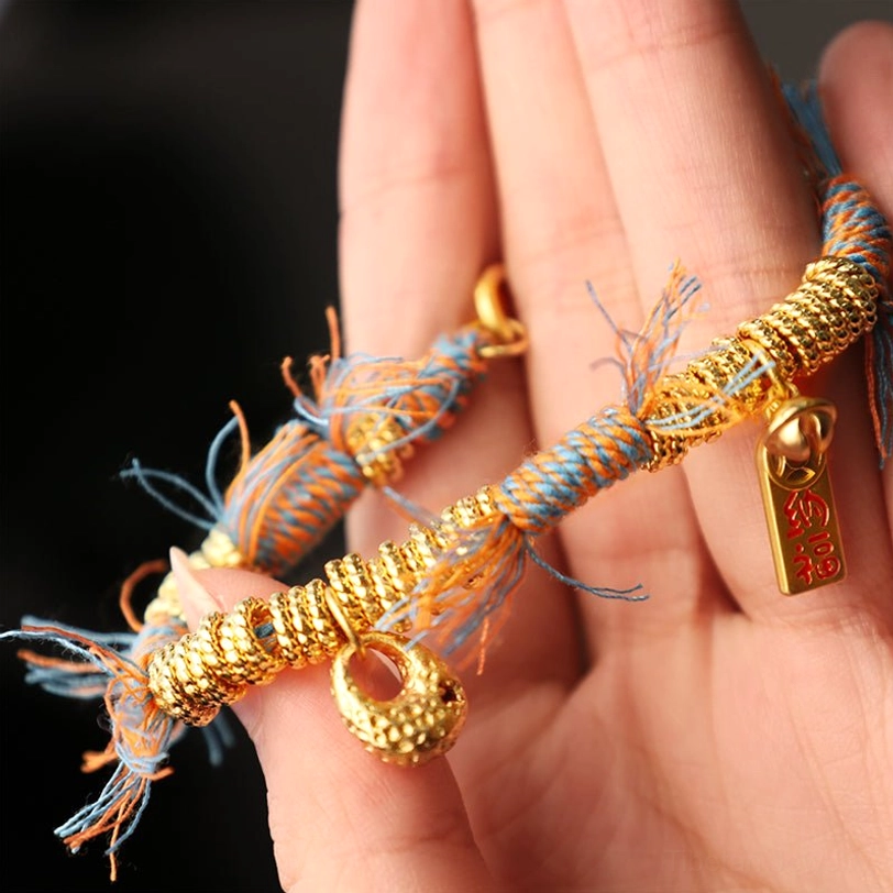Close-up detail shot of handmade Tibetan braided rope bracelet held between fingertips against black background, highlighting blessing charm and gold hardware