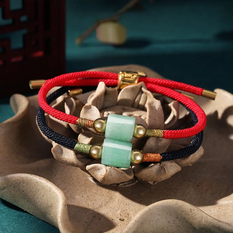 Close-up of woven Tibetan lucky bracelets featuring dual green barrel gemstones, red and black ropes, and gold metal accents on ceramic lotus background