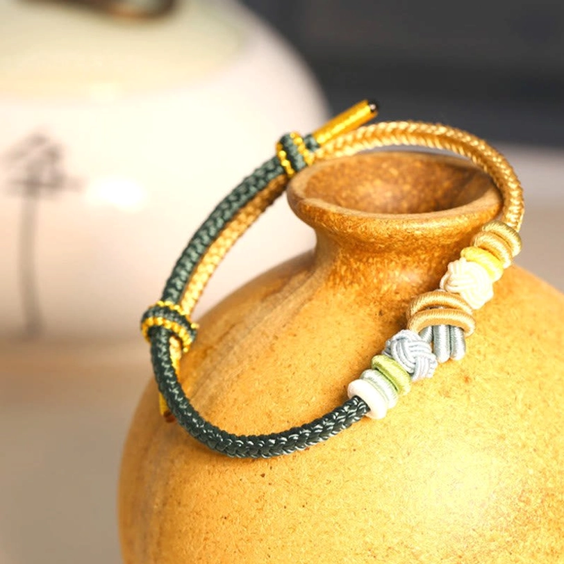 Dark green and gold Tibetan blessed cord bracelet with sacred eight-thread peace knot, displayed with ceramic vase accent