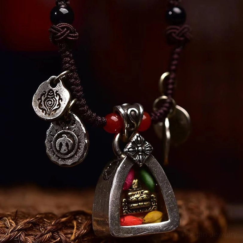 Three-quarter side angle of Tibetan buddha necklace showing braided cord texture, engraved silver charms, and red protective beads on dark fabric background