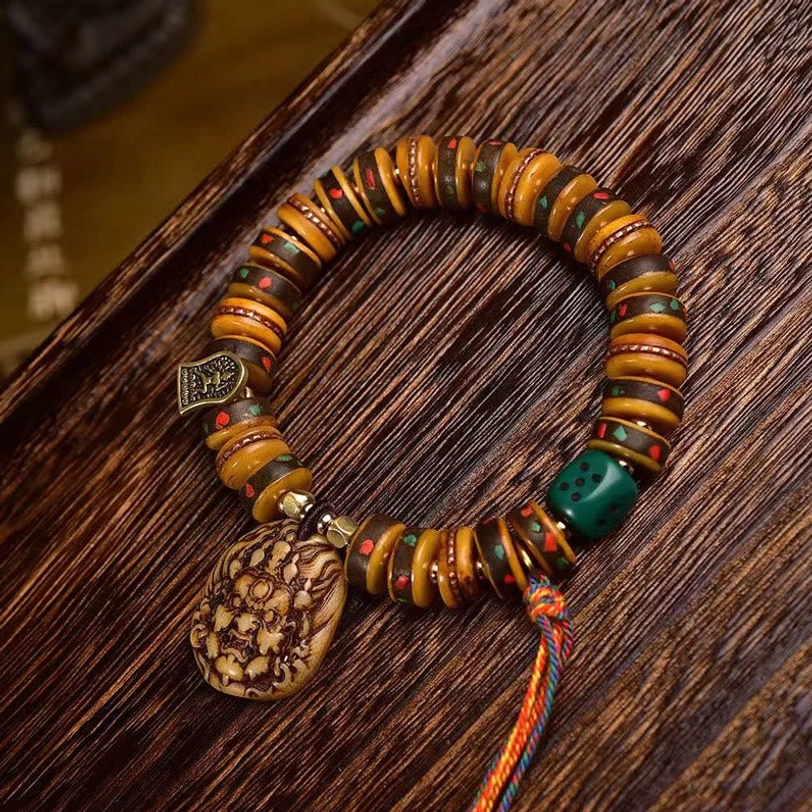 Tibetan camel bone prayer bracelet with folk deity carving, bronze charm, green accent bead and colorful tassel on wooden table, side angle