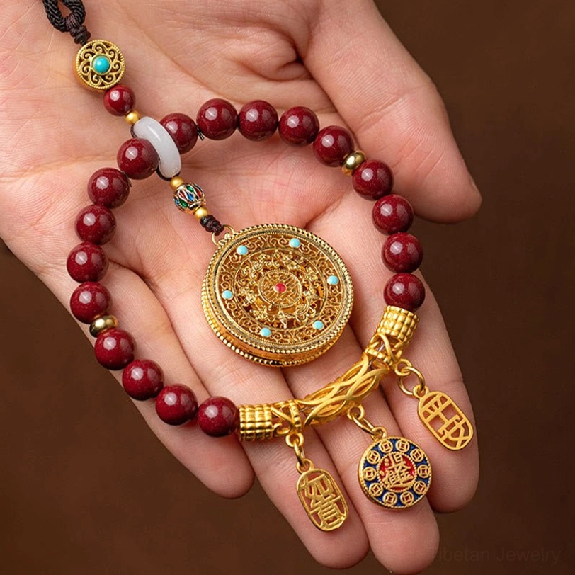 Palmful view of tibetan cinnabar blessing hanging ornament showing maroon bead string and detailed gold filigree round pendant with lucky tags