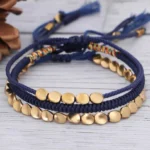 Tibetan copper bead bracelet set displayed overhead on wooden surface with natural pine cone decoration, showing three handwoven styles with dark blue braided cord and gold metal beads with tasseled ends