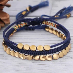 Tibetan copper bead bracelet set displayed overhead on wooden surface with natural pine cone decoration, showing three handwoven styles with dark blue braided cord and gold metal beads with tasseled ends