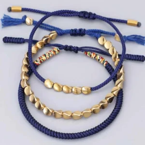 Multi-layer Tibetan copper bead bracelets arranged on white background, featuring dark blue woven cords, mixed gold bead shapes, colorful woven accents, and adjustable tassel ends for layering