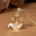 Tibetan copper vajra pendant necklace with dual gold and silver tone, displayed on warm wooden table with blue-brown braided rope showing left three-quarter view and embossed religious relief patterns