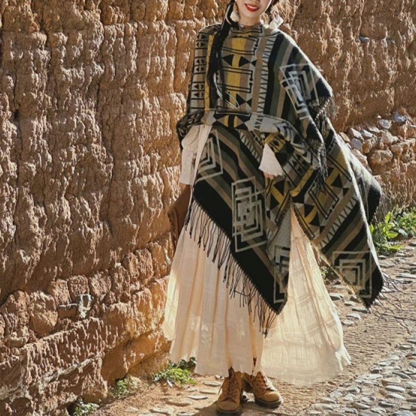 Dark tone ethnic poncho with geometric tribal patterns in green-yellow, tassel hem detail, adobe wall stone alley background