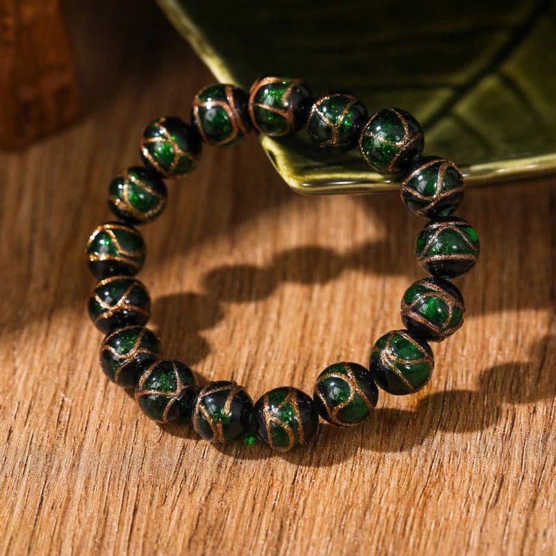 Single dark green Tibetan liuli glass bead bracelet with forest green glazed beads and gold line accents displayed on wooden surface with botanical prop