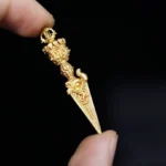 Tibetan Dorje Vajra pendant side view in bright gold tone, held between thumb and index finger against dark background, showing intricate carved relief details