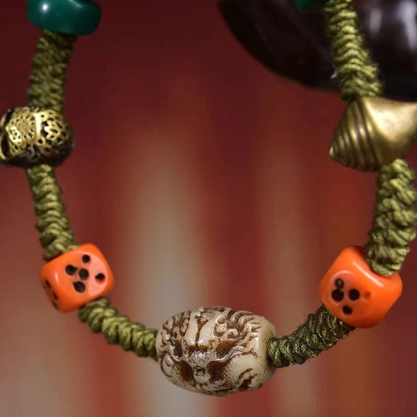 Close-up detail of green braided rope bracelet showing carved dragon bead, orange dice, antique metal skull charm, and stone accents