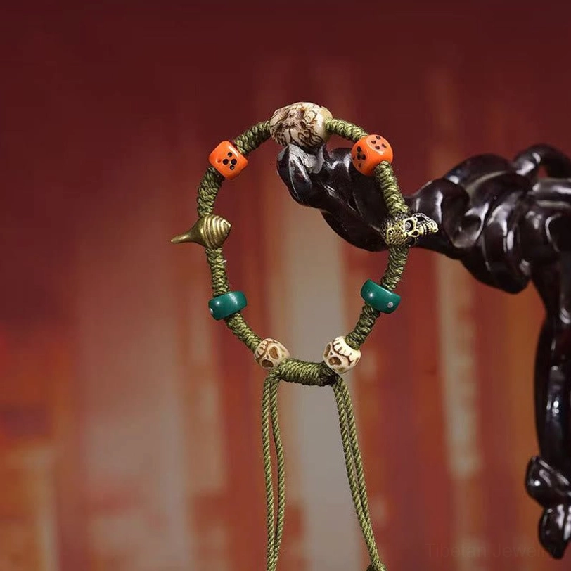 Adjustable Tibetan bracelet with dragon beads, orange dice, skull charms, and green stone hanging from dark wooden prop