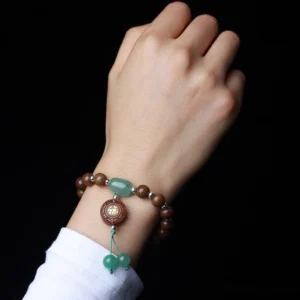 Worn abelia biflora wood protection bracelet on wrist showing green jade stone, fu charm, and tassel beads against white sleeve