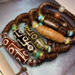 Collection of Tibetan nine-eye dzi bead bracelets in multiple colors including brown, red, black, and green with agate spacers on vintage patterned background