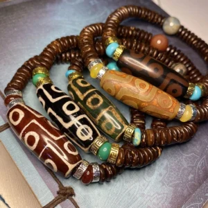 Collection of Tibetan nine-eye dzi bead bracelets in multiple colors including brown, red, black, and green with agate spacers on vintage patterned background