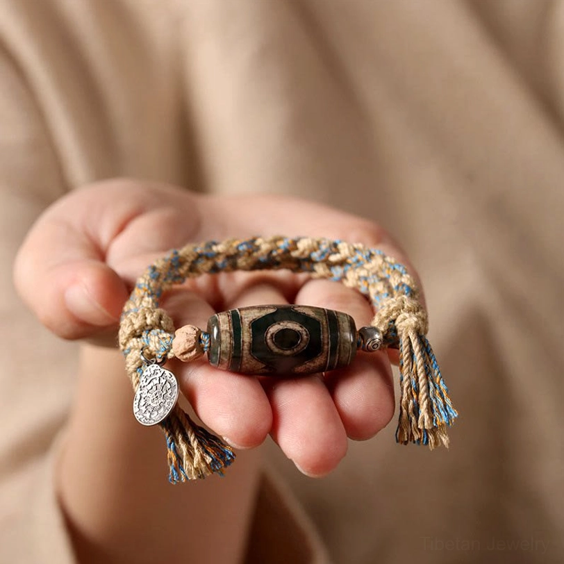 Close-up of Tibetan Dzi bead bracelet held in palm showing detailed craftsmanship of braided cord, sacred stone bead, and tassel accents
