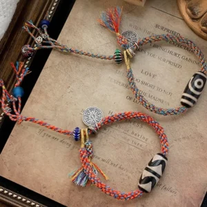 Two handmade Tibetan Dzi bead braided bracelets featuring black-white patterned beads and silver ornaments on wooden tray
