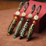 Set of Tibetan Nine-Eye Dzi Bead pendants in multiple colors displayed on brown fabric with red gift box, showing varied eye patterns and braided cord accents