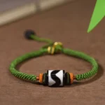Tibetan nine-eye dzi bead bracelet with green braided rope and orange spacer beads, flat lay on wooden table