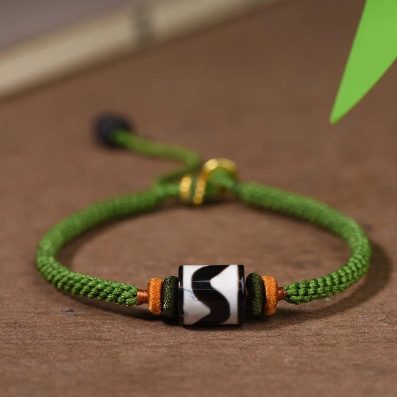 Tibetan nine-eye dzi bead bracelet with green braided rope and orange spacer beads, flat lay on wooden table