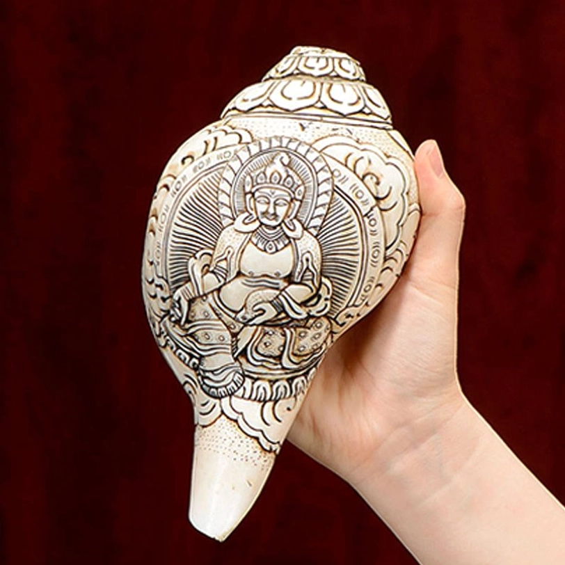 Handheld Tibetan engraved conch shell with seated Buddha figure and halo design on red fabric background