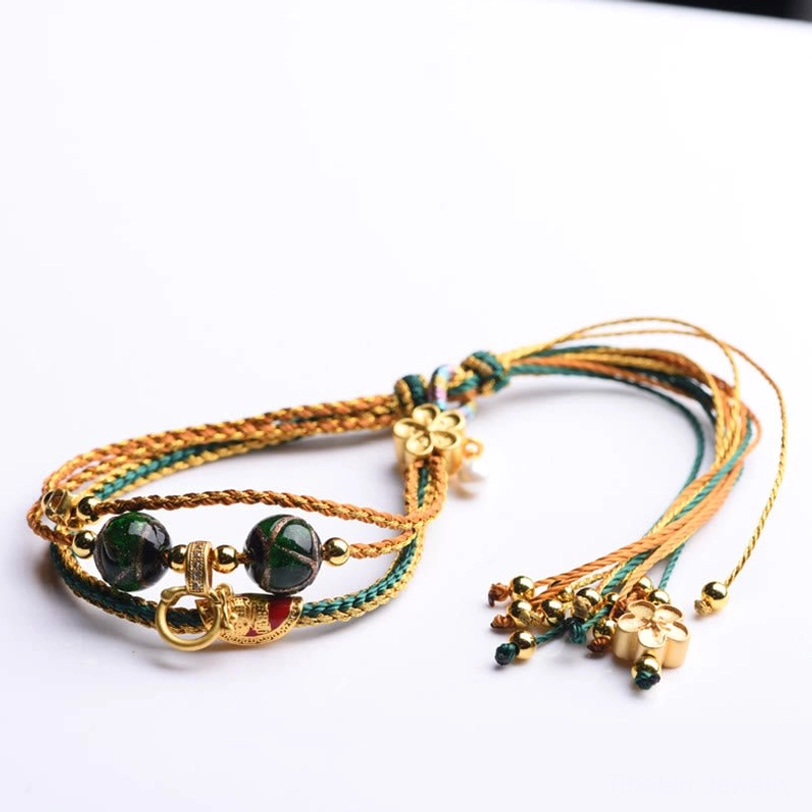 Tibetan handmade braided healing bracelet flat lay on pure white background displaying green gold brown woven ropes dark glass beads and flower tassel pendant