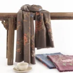 Dark brown Tibetan floral print wrap scarf displayed on wooden bench with vintage teacup and saucer, showcasing soft fuzzy fabric texture and intricate botanical pattern design