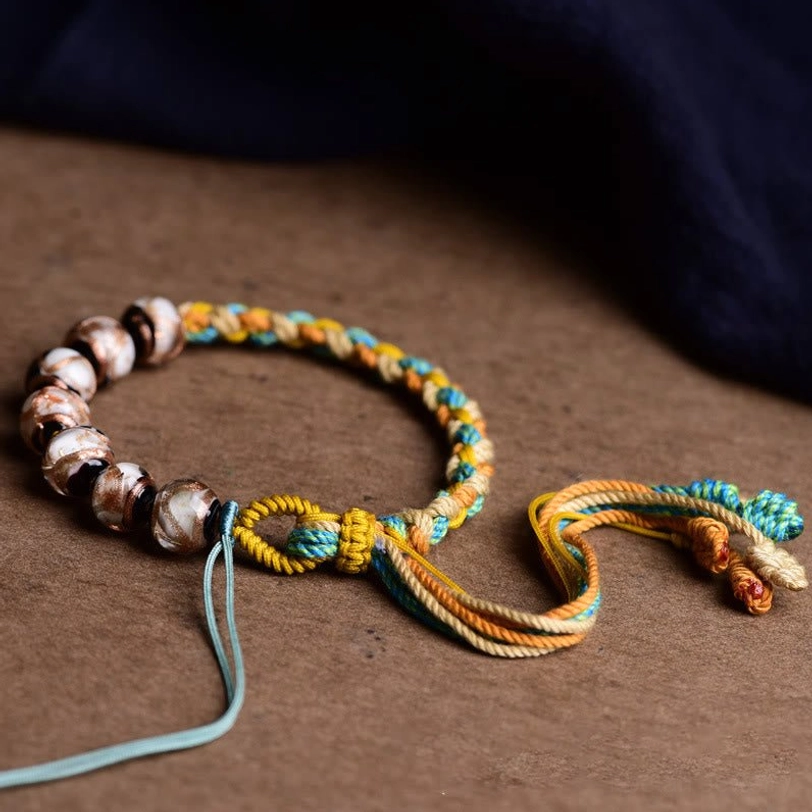 Tibetan liuli glass beaded bracelet with round glazed brown beads, yellow-blue woven rope and blue accent tassel, close-up flat lay side view